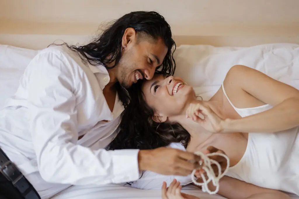 Greta and Jairo lying closely on a bed, smiling at each other while holding a delicate pearl necklace – a tender, intimate moment filled with warmth and connection.